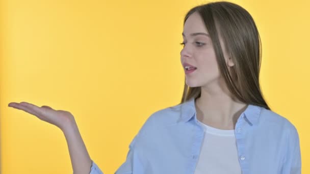 Mujer joven mostrando el producto en la mano, fondo amarillo — Vídeo de stock