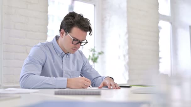 Серьезный молодой человек пишет на бумаге в современном офисе — стоковое видео