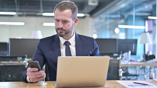 Der junge Geschäftsmann mit Smartphone und Laptop im Büro — Stockfoto