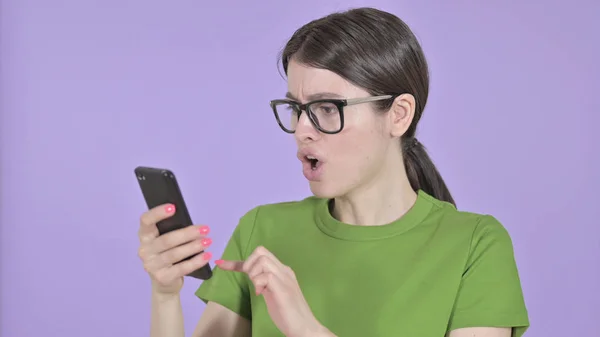 Шокована молода жінка використовує мобільний телефон на фіолетовому тлі — стокове фото