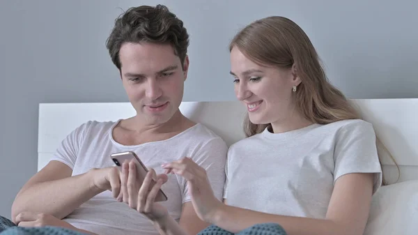 O retrato de casal alegre sentado na cama e usando Smartphone — Fotografia de Stock