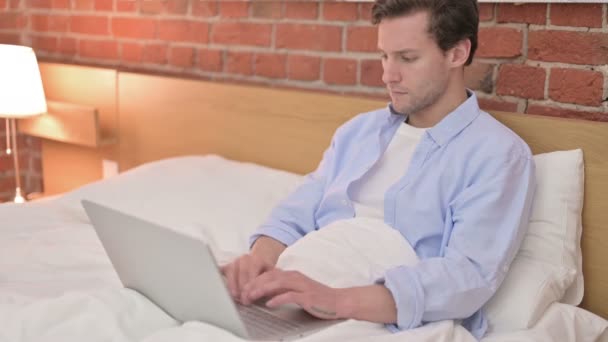 Εστιασμένος νεαρός άνδρας που χρησιμοποιεί φορητό υπολογιστή στο κρεβάτι — Αρχείο Βίντεο