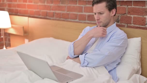 Молодой человек думает и работает на ноутбуке в постели — стоковое видео