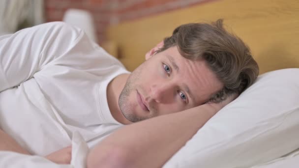 Портрет усталого молодого человека, смотрящего в камеру с кровати — стоковое видео