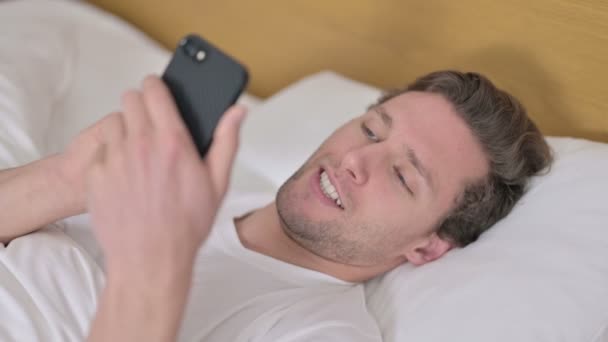 Portrait of Cheerful Young Man using Smartphone in Bed — Stock Video