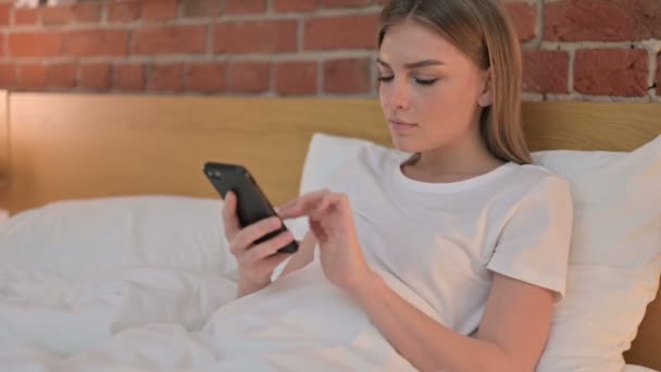 Веселая молодая женщина празднует успех на смартфоне в постели — стоковое видео