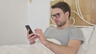 Sakallı genç adam yatakta akıllı telefon kullanıyor.