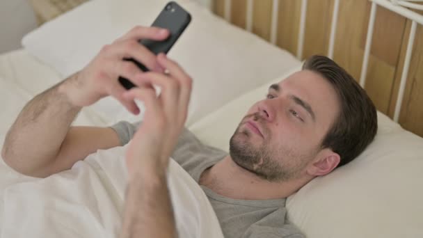 ベアード若い男はベッドでスマートフォンをスクロールします — ストック動画