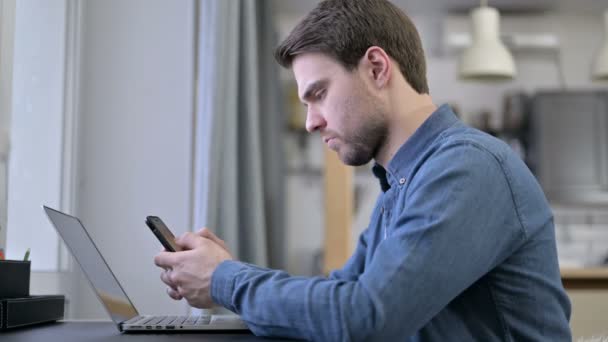 Sakallı genç adam akıllı telefon ve dizüstü bilgisayar kullanıyor. — Stok video