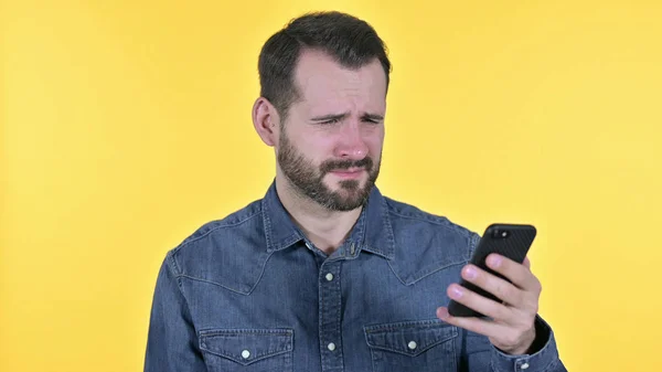 Портрет молодого человека, реагирующего на потерю на смартфоне, желтый фон — стоковое фото