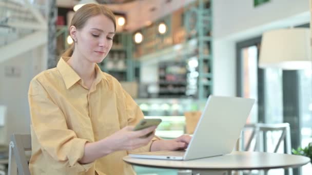 Молодая женщина с ноутбуком разговаривает на смартфоне в кафе — стоковое видео