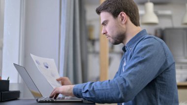Young Man Entering Data from Documents to Laptop