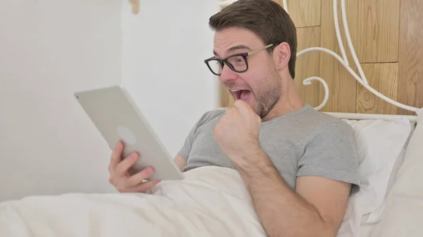 Excited Young Man Surprised by Results on Tablet in bed — 스톡 사진