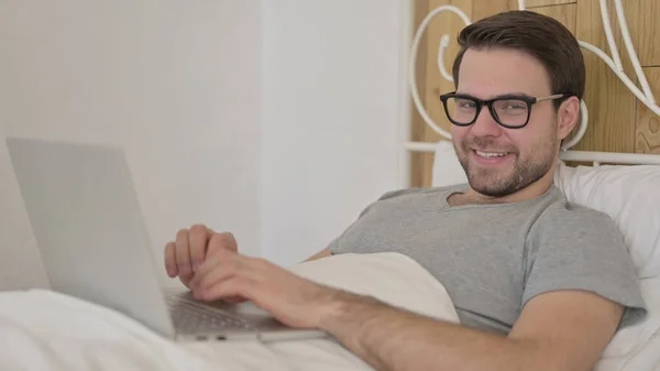 Hombre joven en la cama sonriendo en la cámara, usando el ordenador portátil —  Fotos de Stock