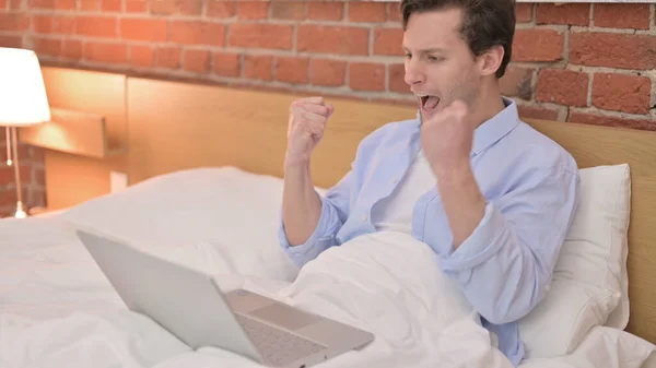 Молодий чоловік святкує успіх на ноутбуці в ліжку — стокове фото