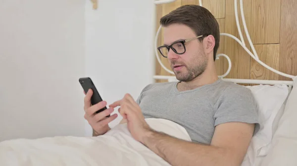 Hombre joven usando Smartphone en la cama —  Fotos de Stock