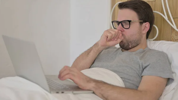 Homem novo que tem tosse ao usar o portátil na cama — Fotografia de Stock
