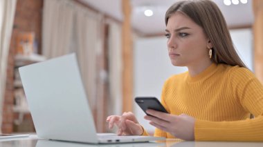 Akıllı telefon kullanan ve dizüstü bilgisayarda çalışan genç bir kadın.