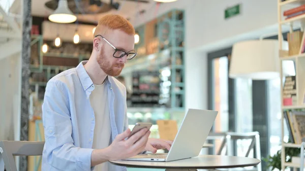 Redhead Man using Smartphone and Laptop in Cafe