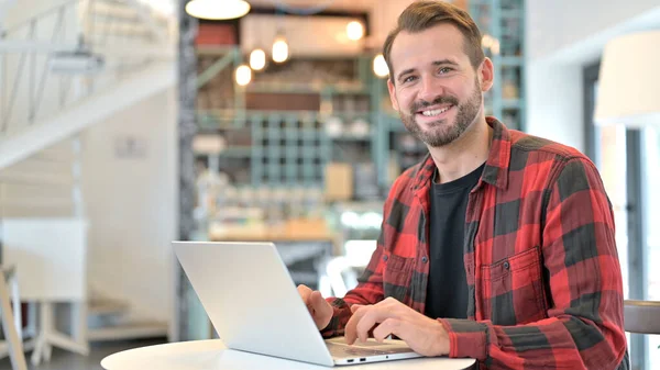 Бородатый молодой человек с ноутбуком улыбается перед камерой в кафе — стоковое фото