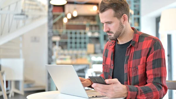 Laptoplu Sakallı Genç Adam Cafe 'de akıllı telefon kullanıyor. — Stok fotoğraf