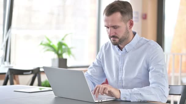 Молодой человек с ноутбуком смотрит в камеру в офисе — стоковое видео