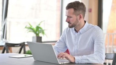 Dizüstü bilgisayarlı genç adam ofiste kameraya gülümsüyor.