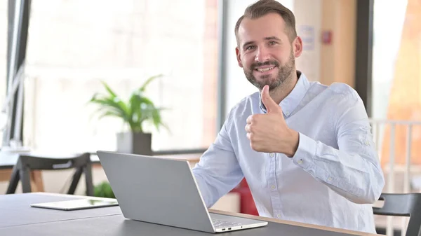 Молодой человек с ноутбуком делает большие пальцы в офисе — стоковое фото