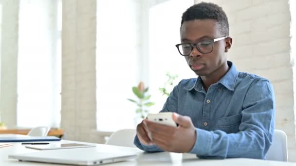 Задоволена молода африканська людина реагує на втрату через смартфон в офісі — стокове відео