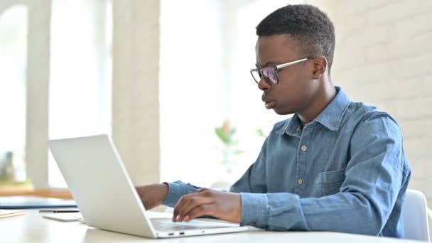 Joven hombre africano con el ordenador portátil enojado en el teléfono en la oficina — Vídeos de Stock