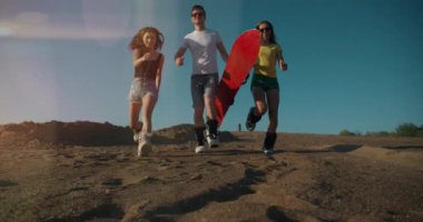 Yaz yaşam tarzı. Kırmızı snowboardlu üç genç koşuyor ve kameraya kum atıyor. Tasasız gençler birlikte eğleniyorlar. Özgürlük, spor, ekstrem, kum kaykayı, seyahat, turizm, tatil konsepti.