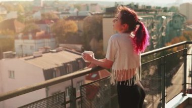 Mutlu esmer kadın balkonda bir fincan çay ya da kahveyle güzel bir günbatımının ya da güneşin doğuşunun ve eski şehrin arka planında dinleniyor. Karantina hayatı. Salgın, koronavirüs, COVID-19 konsepti.