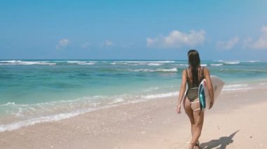 Okyanus sörfü. Mavi deniz sahilinde sörf tahtası ile suya doğru koşan güzel bir sörfçü kız. Güneş parlıyor. Egzotik adada yaz tatili. Spor aktivitesi ve seyahat konsepti.