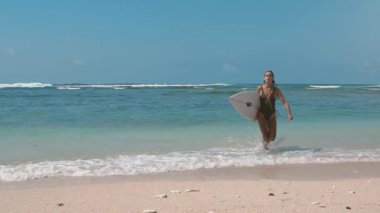 Egzotik adada yaz tatili. Spor aktivitesi ve seyahat konsepti. Okyanus sörfü. Mavi deniz sahilinde sörf tahtası ile suya doğru koşan güzel bir sörfçü kız. Güneş parlıyor.