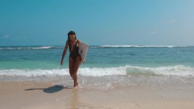 Okyanus sörfü. Mavi deniz sahilinde sörf tahtası ile suya doğru koşan güzel bir sörfçü kız. Güneş parlıyor. Egzotik adada yaz tatili. Spor aktivitesi ve seyahat konsepti.