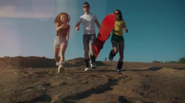 Yaz yaşam tarzı. Kırmızı snowboardlu üç genç koşuyor ve kameraya kum atıyor. Tasasız gençler birlikte eğleniyorlar. Özgürlük, spor, ekstrem, kum kaykayı, seyahat, turizm, tatil konsepti.