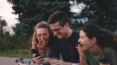 Üç tane çok ırklı genç insan birlikte vakit geçiriyor. Mutlu gülümseyen öğrenciler üniversite binasının önünde gülüyorlar ve cep telefonlarına bakıyorlar. Arkadaşlık, iletişim, gençlik ve yaşam tarzı kavramı