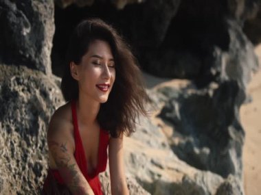 Moda kırmızısı elbiseli, kumsalda rahatlatıcı genç Asyalı esmer kadın. Güzel dişi deniz kenarında kayaların üzerinde oturmaktan mutlu güneş parlarken, rüzgar saçlarını dalgalandırırken. Yaşam tarzı, seyahat ve tatil konsepti.