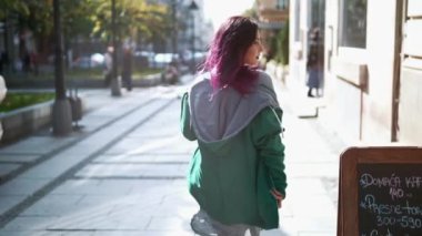 Kamerada flört eden gülümseyen kızı kapat. Güzel bir kadın eski şehir sokağına gidiyor ve güneş parlıyor. Mor saçlı, güneşli caddede yürüyen seksi ve şık bir kadının arka planı..