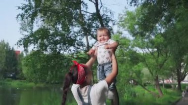 Mutlu, genç bir anne, küçük sevimli oğlunu ellerinin arasında tutuyor ve parkta gülüyor. Yeşil bahçede bebekle birlikte yürüyen bir kadın. Aile kavramı, çocukluk, mutluluk, ebeveynlik.