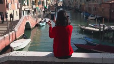 Kırmızı kazaklı güzel Asyalı kızın köprünün kenarından akıllı telefonuyla Venedik Kanalı 'nın fotoğrafını çekişi. Hoş bir kadın tatilini Venedik 'te geçiriyor..