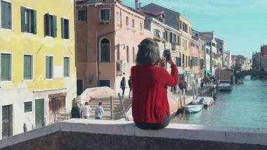 İtalya 'da güneşin sıcacık ışınlarında dinlenmek. Köprünün kenarında oturan kırmızı kazaklı çekici esmer kızın arkadan görünüşü süzülen gondollara ve kanal kenarında yürüyen turistlere bakıyor..