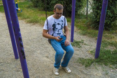 Bir zincir adam salıncağa salıncak ve sörf Internet aracılığıyla bir smartphone. Genç adam, atlıkarınca üstünde haddeleme ve tablet ile sohbette yeniden aittir. Bir adam fetbolke ve kot pantolon.