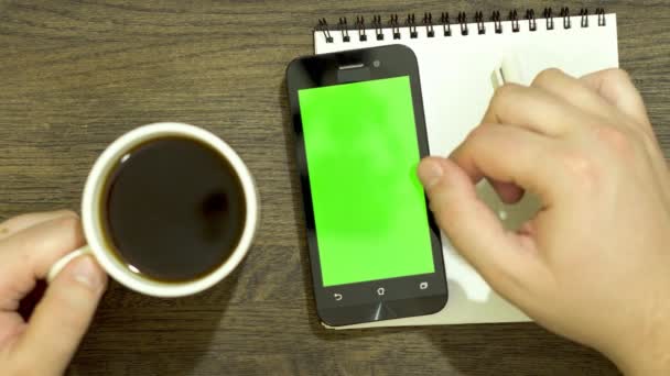 Primer plano de una mano trabajando en un smartphone. Fondo de madera. Sobre la mesa hay un cuaderno. La otra mano toma una taza de café . — Vídeo de stock