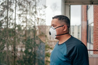 Balkonda koruyucu maskeli İspanyol bir adam var. 2020 pandemik koronavirüs. Karantina. Virüs konsepti. Salgın hastalık. Koronavirüs salgını yüzünden karantina sırasında balkondan dışarı bakan maskeli genç bir İspanyol. Üzgün bir adamın portresi.