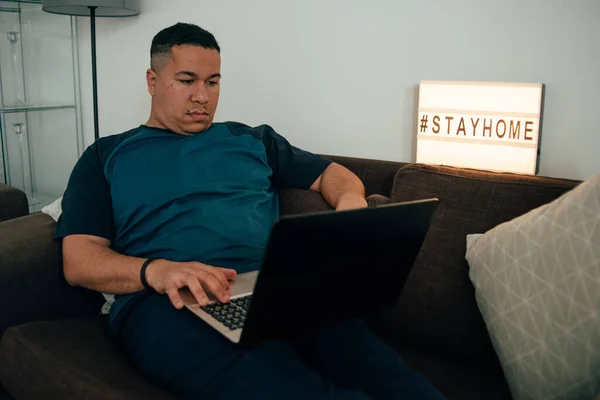 Genç İspanyol adam karantina sırasında kanepede uzanıyor ve dizüstü bilgisayarının başında çalışıyor. İnsan, başkalarının tehlikelerinden kaçınmak için evden çalışır. Genç İspanyol adam koronavirüsün yayılmasını nasıl durduracağını düşünüyor. Etiketi evde kal. hashtag Evde kal.
