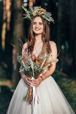 Şık bir buket ve çelengi olan muhteşem, mutlu esmer gelin portresi. Şık bir gelin ormanın arka planında buketle poz veriyor, ormanın dışında lüks, görkemli bir boho düğünü. Doğal makyaj, saç stili, çelenk..