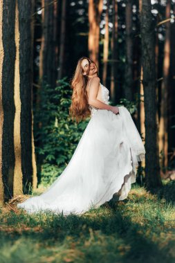 Şık bir gelin ormanın arka planında poz veriyor ve gülümsüyor. Ormanda lüks, muhteşem bir boho düğünü. Beyaz elbiseli mutlu gelin arka planda güzel orman manzaralı.