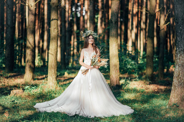 Beautiful fabulous happy brunette bride with stylish bouquet and wreath. stylish bride posing with bouquet on background of forest, luxury gorgeous boho wedding at forest outdoors.