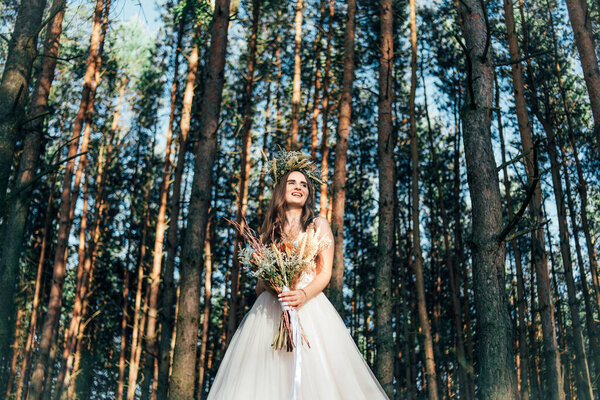 Happy bride in white stylish dress, with a wedding bouquet, beautiful forest landscape in the background. Natural makeup, hairstyle, wreath. bride in the forest with a bouquet of dried flowers